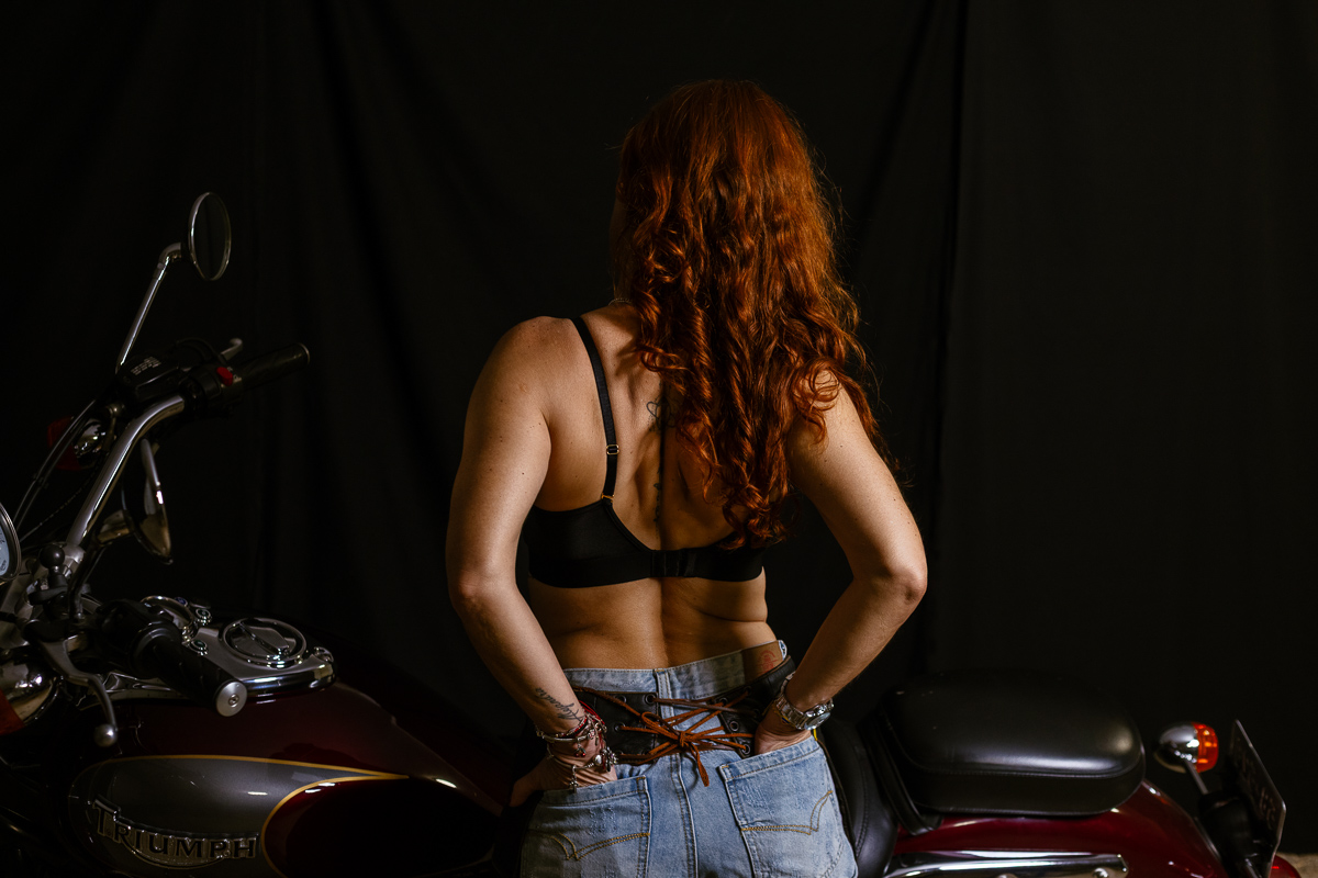 Beautiful redhead in jeans shorts and a black bra standing before a motorcycle with her back turned to the camera